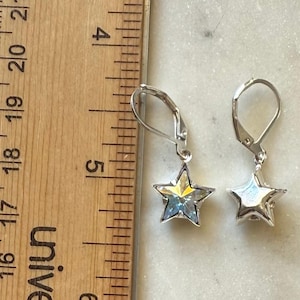 Puede incluir: Un par de pendientes plateados en forma de estrella con cierres de palanca. Los pendientes tienen un centro facetado e iridiscente y se muestran junto a una regla de madera. Los pendientes miden aproximadamente 1,3 cm de largo.