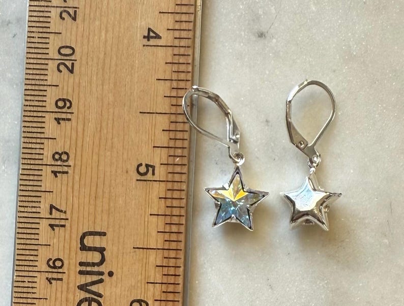 Puede incluir: Un par de pendientes plateados en forma de estrella con cierres de palanca. Los pendientes tienen un centro facetado e iridiscente y se muestran junto a una regla de madera. Los pendientes miden aproximadamente 1,3 cm de largo.