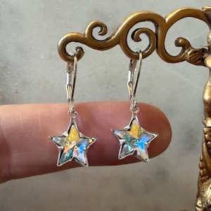 Puede incluir: Un par de pendientes en forma de estrella con marco plateado. Las estrellas tienen un centro amarillo y facetas iridiscentes que reflejan tonos azules, verdes y morados. Los pendientes cuelgan de cierres de palanca.