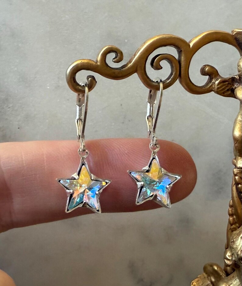 Puede incluir: Un par de pendientes en forma de estrella con marco plateado. Las estrellas tienen un centro amarillo y facetas iridiscentes que reflejan tonos azules, verdes y morados. Los pendientes cuelgan de cierres de palanca.