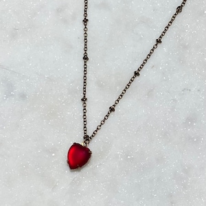 May include: A delicate gold chain necklace with a red glass heart pendant. The chain features small beads.