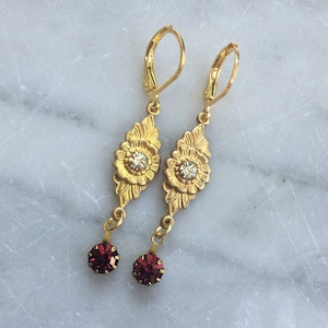 Op de afbeelding: Gouden oorhangers met een bloemmotief en een heldere strasssteen in het midden. De oorhangers hebben een kleine rode strasssteen die aan de onderkant hangt.
