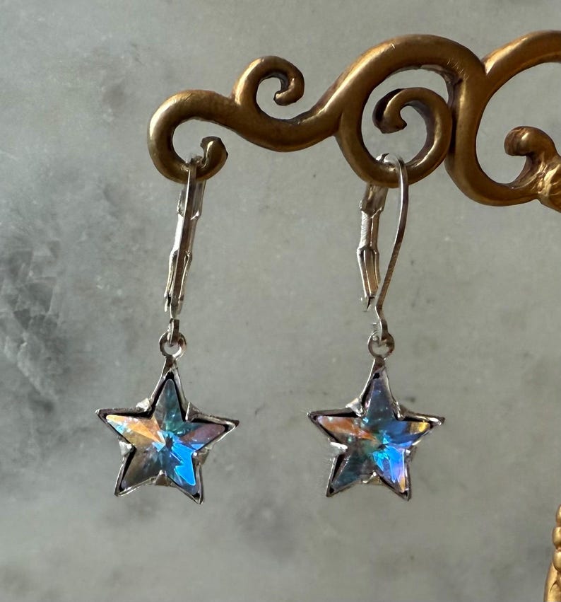 Puede incluir: Un par de pendientes plateados en forma de estrella. Cada pendiente presenta una estrella de cinco puntas con facetas iridiscentes multicolores. Los pendientes cuelgan de cierres de palanca.