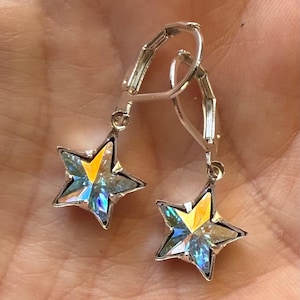 Puede incluir: Un par de pendientes plateados en forma de estrella. Cada pendiente presenta una estrella de cinco puntas con un centro transparente e iridiscente que refleja tonos amarillos, azules y verdes. Los pendientes tienen un cierre de palanca.