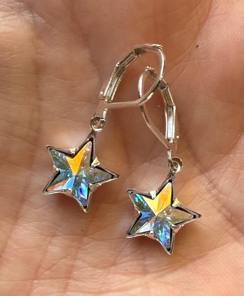 Puede incluir: Un par de pendientes plateados en forma de estrella. Cada pendiente presenta una estrella de cinco puntas con un centro transparente e iridiscente que refleja tonos amarillos, azules y verdes. Los pendientes tienen un cierre de palanca.