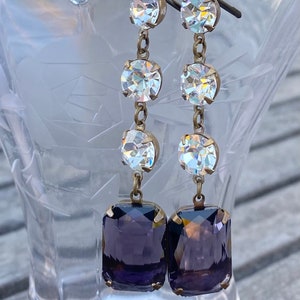 May include: A pair of dangle earrings with a purple square-shaped stone at the bottom and three clear round stones above it.