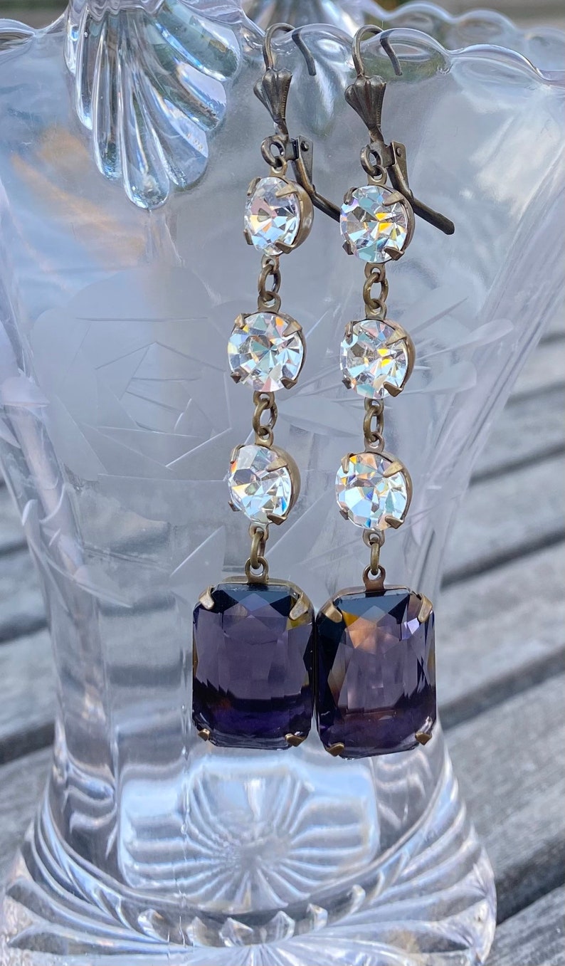May include: A pair of dangle earrings with a purple square-shaped stone at the bottom and three clear round stones above it.