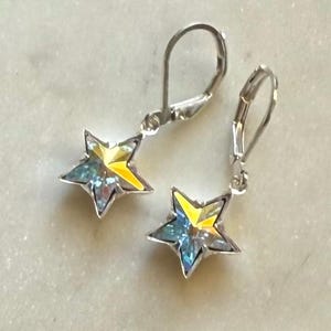 Puede incluir: Un par de pendientes de plata con cierre de palanca y colgantes en forma de estrella. Los colgantes de estrella tienen un dise&ntilde;o de cristal iridiscente amarillo y azul. Los pendientes se muestran sobre una superficie clara y texturizada.