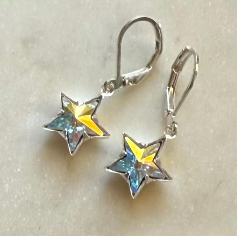 Puede incluir: Un par de pendientes de plata con cierre de palanca y colgantes en forma de estrella. Los colgantes de estrella tienen un dise&ntilde;o de cristal iridiscente amarillo y azul. Los pendientes se muestran sobre una superficie clara y texturizada.