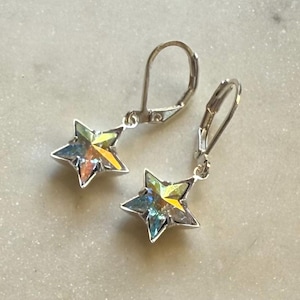 Puede incluir: Un par de pendientes plateados con forma de estrella. Cada pendiente presenta una estrella de cinco puntas con cristales iridiscentes multicolores. Los pendientes tienen cierres de palanca.