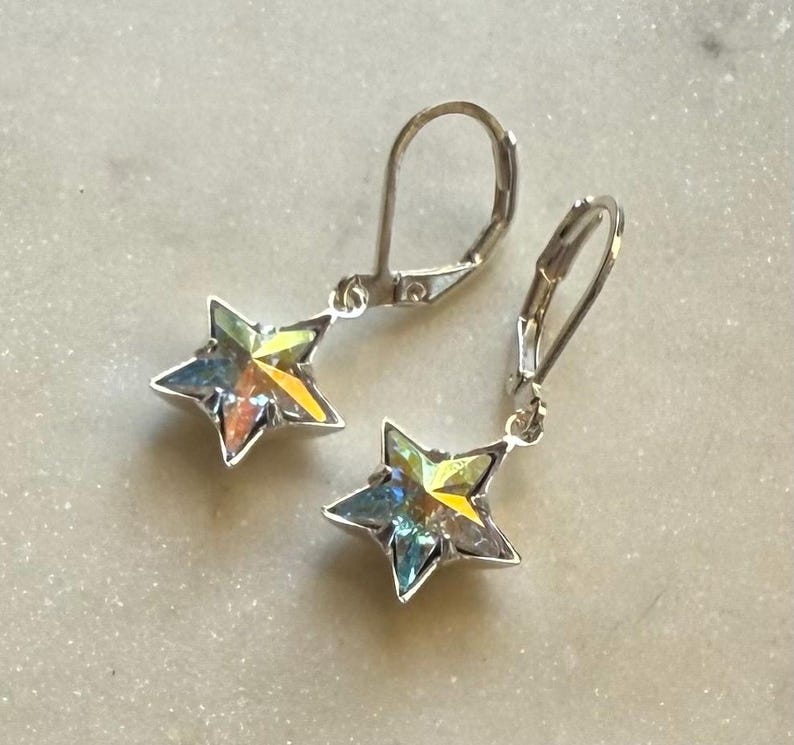 Puede incluir: Un par de pendientes plateados con forma de estrella. Cada pendiente presenta una estrella de cinco puntas con cristales iridiscentes multicolores. Los pendientes tienen cierres de palanca.