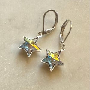 Puede incluir: Un par de pendientes plateados con cierre de palanca. Cada pendiente presenta un colgante de estrella de cinco puntas con un acabado brillante e iridiscente, que muestra tonos de amarillo, azul y verde. Los pendientes est&aacute;n dise&ntilde;ados para orejas perforadas.