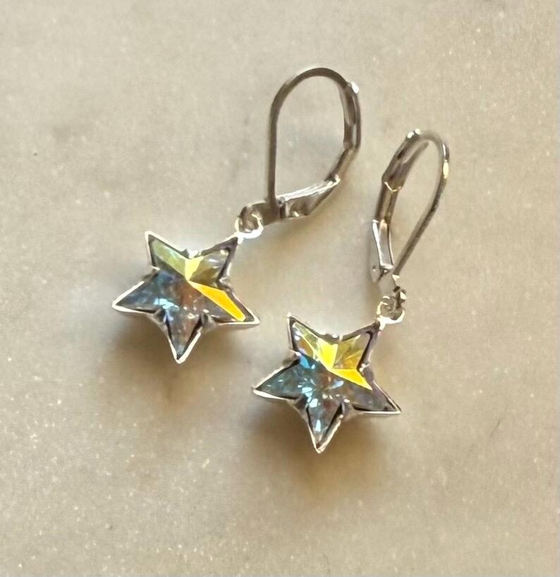 Puede incluir: Un par de pendientes plateados con cierre de palanca. Cada pendiente presenta un colgante de estrella de cinco puntas con un acabado brillante e iridiscente, que muestra tonos de amarillo, azul y verde. Los pendientes est&aacute;n dise&ntilde;ados para orejas perforadas.