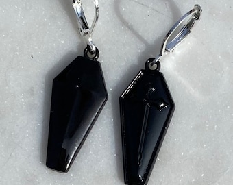 May include: A pair of black coffin-shaped earrings with silver clip-on backs. The earrings feature a raised design of a dagger inside the coffin.