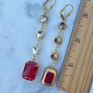 May include: A pair of gold earrings with a red and clear crystal design. The earrings are approximately 2 inches long and feature a red rectangular crystal at the bottom.