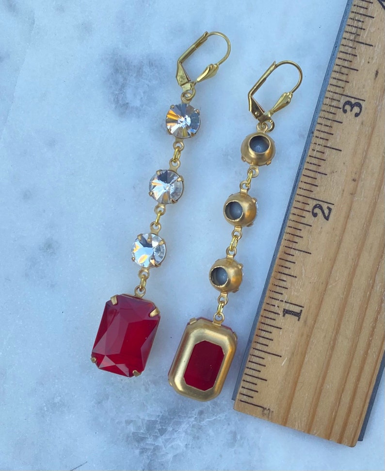 May include: A pair of gold earrings with a red and clear crystal design. The earrings are approximately 2 inches long and feature a red rectangular crystal at the bottom.