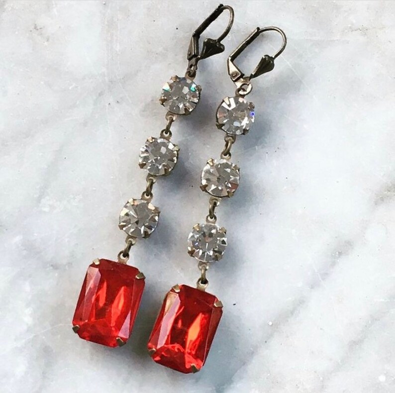 May include: A pair of long, dangling earrings with a red rectangular stone at the bottom and four clear round stones above it.