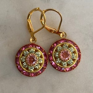 May include: A pair of gold dangle earrings with pink and white crystal accents. The earrings feature a round design with a central pink crystal surrounded by smaller white crystals.
