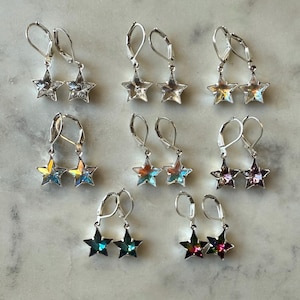 Puede incluir: Una colecci&oacute;n de pendientes en forma de estrella en varios colores, incluyendo transparente, iridiscente y morado. Los pendientes tienen un cierre de palanca y se muestran sobre una superficie clara. Los pendientes son probablemente de metal y cristal.
