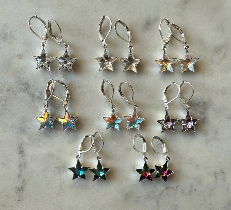Puede incluir: Una colecci&oacute;n de pendientes en forma de estrella en varios colores, incluyendo transparente, iridiscente y morado. Los pendientes tienen un cierre de palanca y se muestran sobre una superficie clara. Los pendientes son probablemente de metal y cristal.