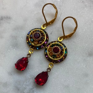 May include: A pair of gold dangle earrings with a red teardrop-shaped crystal dangling from each earring. The top of each earring features a round, multi-colored crystal design.