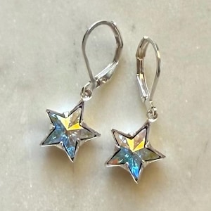 Puede incluir: Un par de pendientes plateados en forma de estrella. Cada pendiente presenta una estrella de cinco puntas con un centro transparente e iridiscente que refleja tonos amarillos, azules y verdes. Los pendientes tienen cierres de palanca.