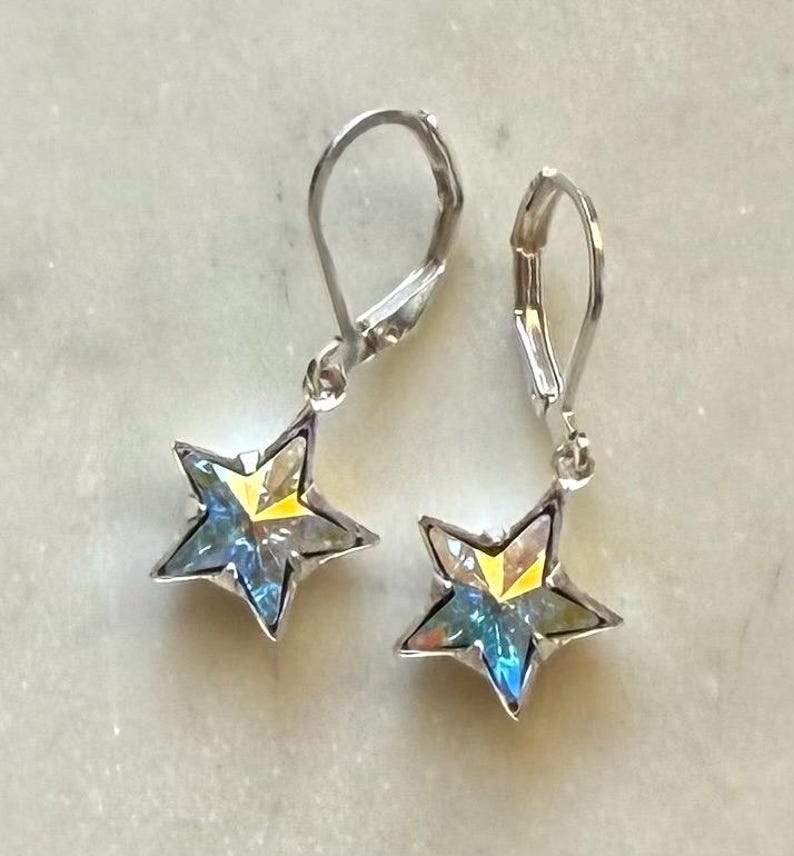 Puede incluir: Un par de pendientes plateados en forma de estrella. Cada pendiente presenta una estrella de cinco puntas con un centro transparente e iridiscente que refleja tonos amarillos, azules y verdes. Los pendientes tienen cierres de palanca.
