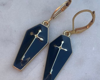 May include: A pair of gold-tone earrings with a blue coffin design and a gold cross. The earrings have a hook closure.