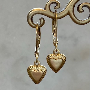 Peut inclure: Une paire de boucles d'oreilles dorées en forme de cœur. Chaque boucle d'oreille présente un pendentif en forme de cœur avec une bordure texturée et une fermeture à levier. Les boucles d'oreilles sont présentées sur un support décoratif doré.