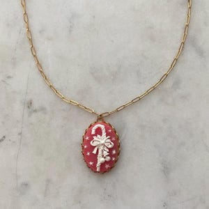 May include: A gold chain necklace with a pink cameo pendant featuring a white candy cane with a bow and bells.
