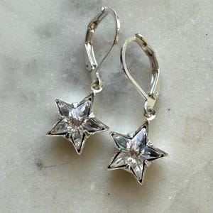 May include: A pair of silver leverback earrings. Each earring has a clear, faceted star-shaped charm. The earrings are shown on a light-coloured, marbled surface.