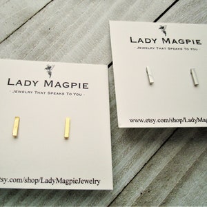 May include: Two pairs of minimalist bar earrings on display cards. The earrings are made of gold and silver metal. The cards have the text "Lady Magpie - Jewelry that speaks to you" and a website address.