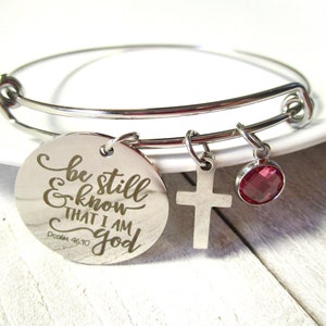 May include: A silver-colored bangle bracelet with three charms. One charm is a round disc with the inscription "be still & know THAT I AM GOD" and a cross. A red gemstone charm hangs from the bracelet.