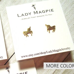 Puede incluir: Pendientes de bot&oacute;n de unicornio dorados en una tarjeta blanca con el texto "Lady Magpie - Joyer&iacute;a que te habla." y la direcci&oacute;n del sitio web "www.etsy.com/shop/LadyMagpieJewelry".