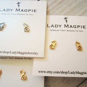 May include: Cat-shaped stud earrings in silver, gold, and rose gold. The earrings are presented on white cards with the text "LADY MAGPIE JEWELRY THAT SPEAKS TO YOU" and the Etsy shop URL. The cat design is a simple outline with a curled tail.