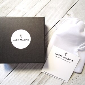 Puede incluir: Una caja negra con un c&iacute;rculo blanco que contiene el texto "Lady Magpie" y una peque&ntilde;a imagen de un hada. La caja est&aacute; sobre una superficie de madera blanca. Una bolsa blanca con cord&oacute;n est&aacute; al lado de la caja. La bolsa tiene una etiqueta blanca adjunta con el texto "Lady Magpie - Joyer&iacute;a que te habla." y una peque&ntilde;a imagen de un hada. La etiqueta tambi&eacute;n tiene la direcci&oacute;n del sitio web "www.etsy.com/shop/LadyMagpieJewelry".