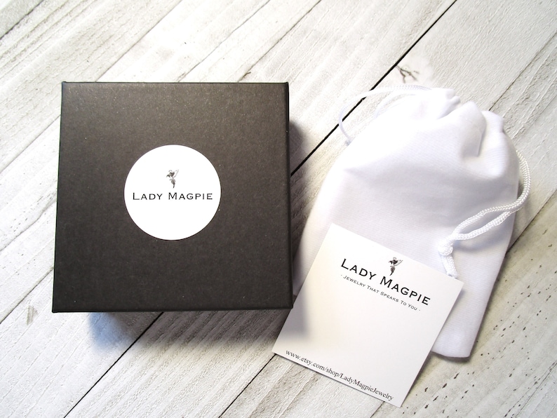 Puede incluir: Una caja negra con un c&iacute;rculo blanco que contiene el texto "Lady Magpie" y una peque&ntilde;a imagen de un hada. La caja est&aacute; sobre una superficie de madera blanca. Una bolsa blanca con cord&oacute;n est&aacute; al lado de la caja. La bolsa tiene una etiqueta blanca adjunta con el texto "Lady Magpie - Joyer&iacute;a que te habla." y una peque&ntilde;a imagen de un hada. La etiqueta tambi&eacute;n tiene la direcci&oacute;n del sitio web "www.etsy.com/shop/LadyMagpieJewelry".
