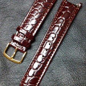 May include: A pair of brown leather watch straps with a crocodile pattern. The straps have a gold buckle and are designed for a watch with a standard lug width.