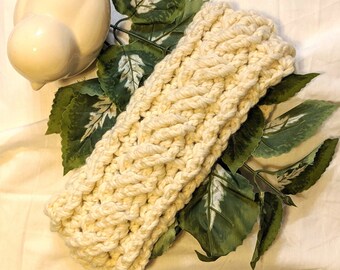 Può includere: Un cerchietto in crochet color crema con un motivo intrecciato. Il cerchietto è appoggiato su una superficie bianca con foglie verdi.