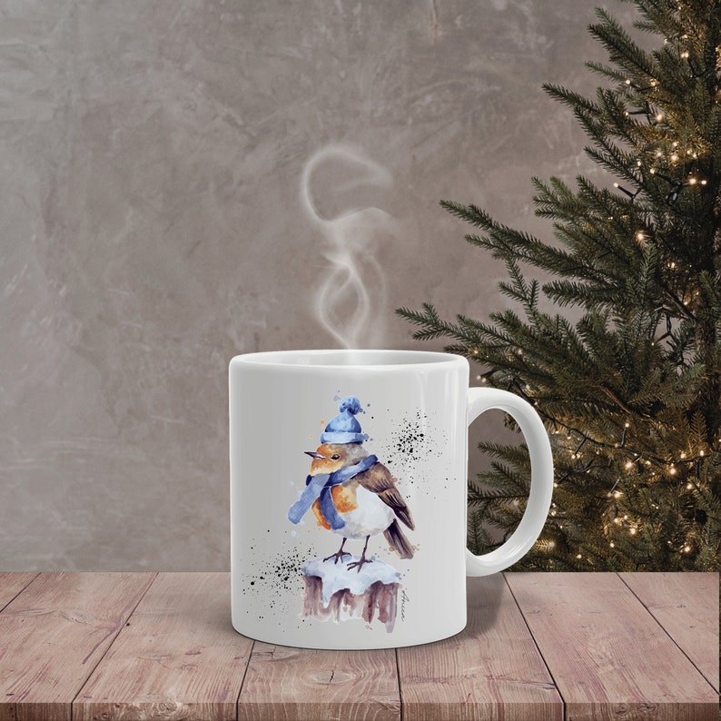 May include: White ceramic mug with a watercolor illustration of a robin wearing a blue hat and scarf, perched on a snow-covered branch. The mug is filled with steam, suggesting a warm beverage.