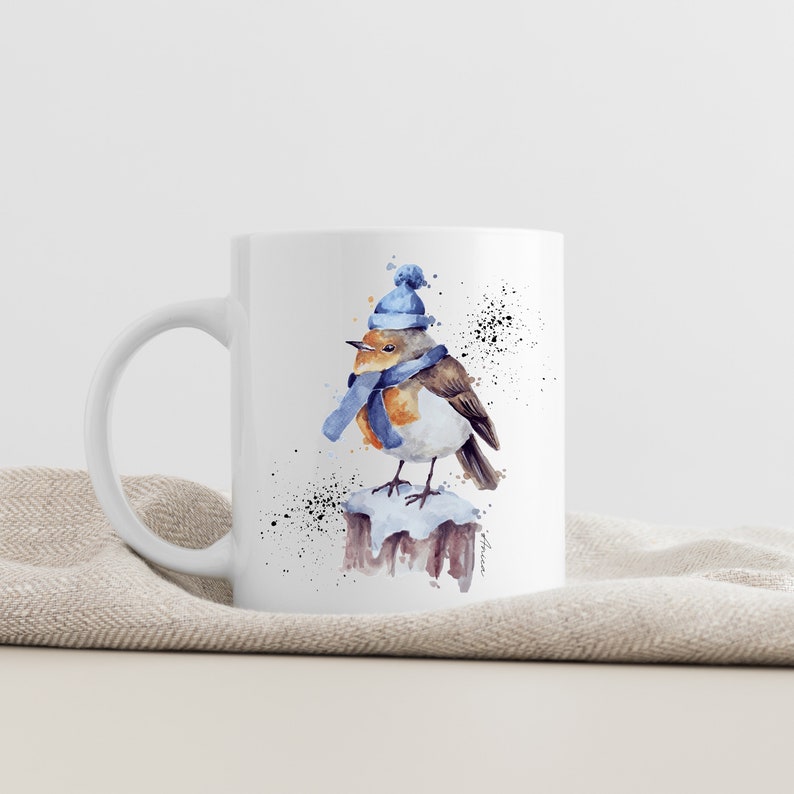May include: White ceramic mug with a watercolor illustration of a robin wearing a blue hat and scarf, perched on a snow-covered branch.