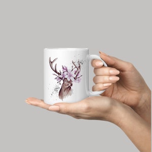 Könnte beinhalten: Weiße Keramiktasse mit einer Aquarell-Illustration eines Hirsches mit einer Blumenkrone aus rosa Blüten. Der Hirsch hat braune Geweihe und ein braunes Gesicht.