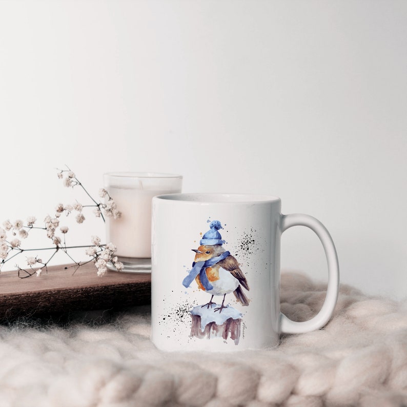 May include: White ceramic mug with a watercolor illustration of a robin wearing a blue hat and scarf. The robin is perched on a snow-covered branch.