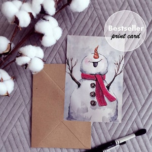 May include: Watercolor painting of a snowman with a red scarf and a playful expression. The snowman is wearing buttons and has a carrot nose. The painting is on a white background and has a brown envelope and paintbrushes in the background. The text "Bestseller print card" is visible in the top right corner of the image.