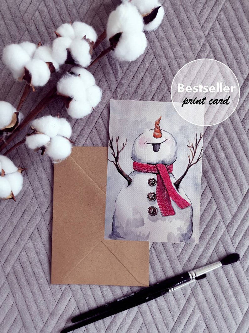 May include: Watercolor painting of a snowman with a red scarf and a playful expression. The snowman is wearing buttons and has a carrot nose. The painting is on a white background and has a brown envelope and paintbrushes in the background. The text "Bestseller print card" is visible in the top right corner of the image.