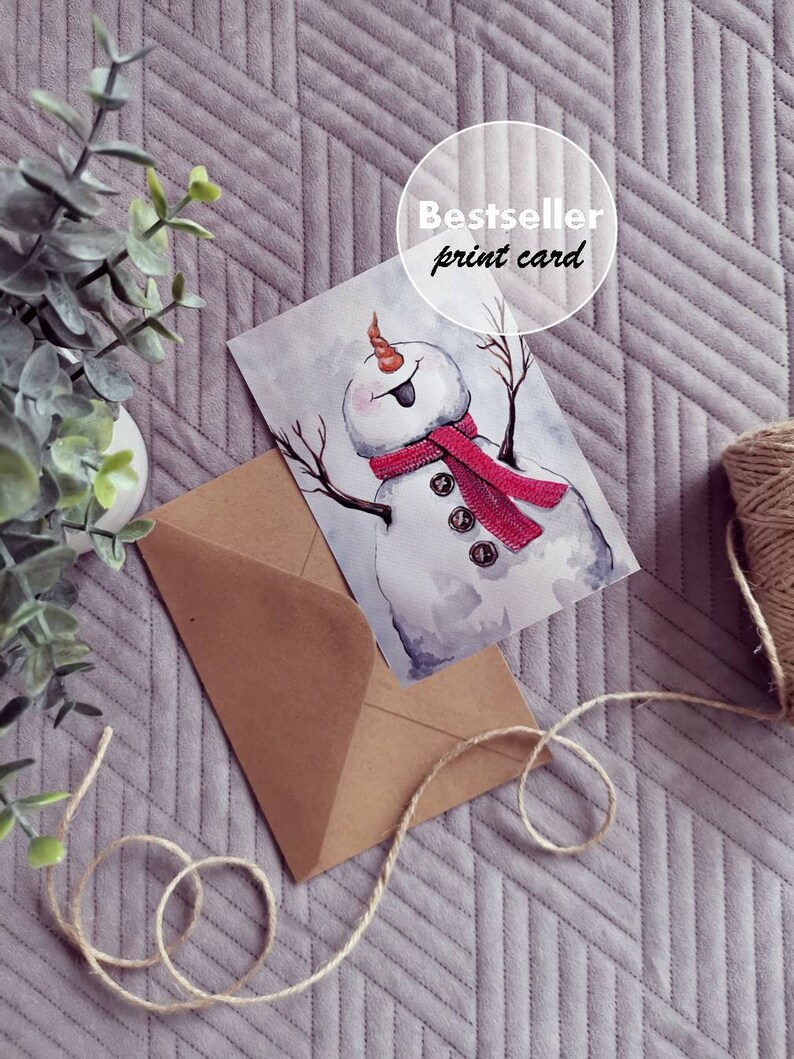 May include: A watercolor illustration of a snowman with a red scarf and buttons. The illustration is printed on a white card with the text "Bestseller print card" in a circle.