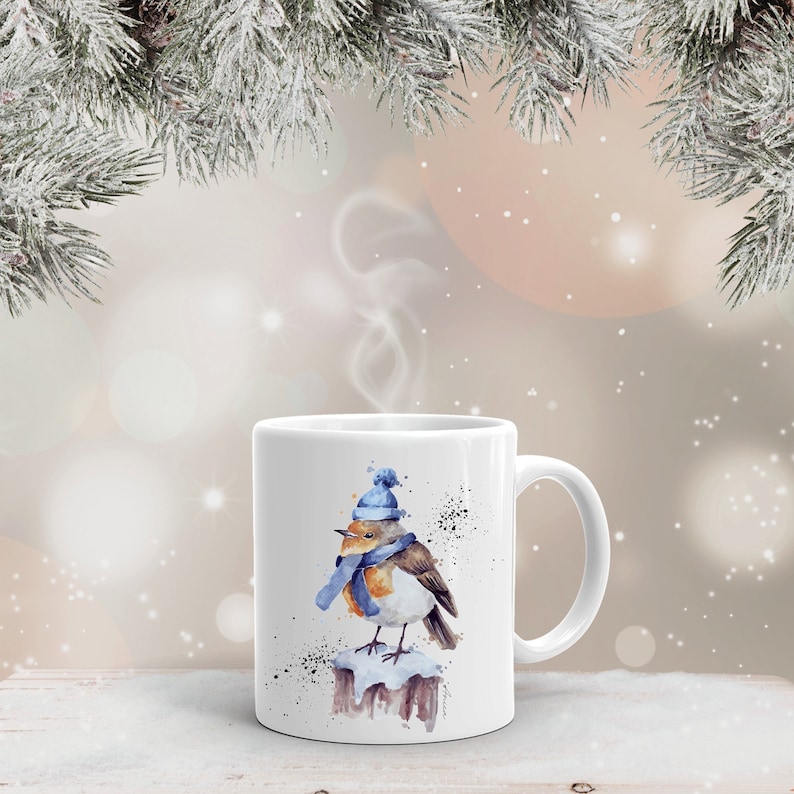 May include: White ceramic mug with a watercolor illustration of a robin wearing a blue hat and scarf, perched on a snow-covered branch. The robin is facing left and has a brown breast and a yellow beak.