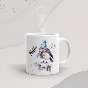 May include: White ceramic mug with a watercolor illustration of a robin wearing a blue hat and scarf. The mug has the name "Stella" written on it in black cursive.