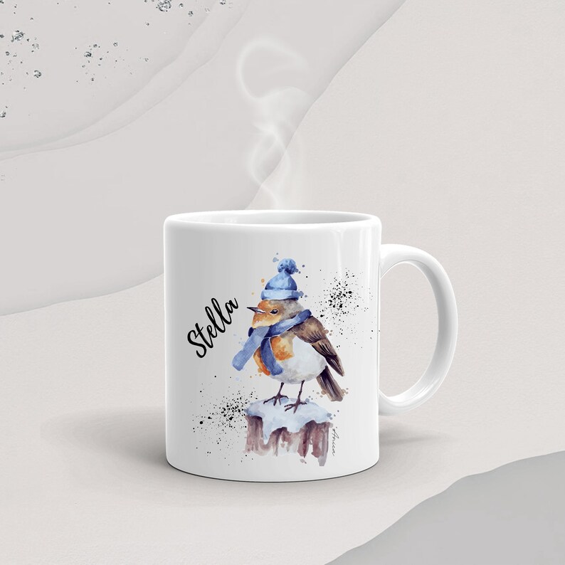 May include: White ceramic mug with a watercolor illustration of a robin wearing a blue hat and scarf. The mug has the name "Stella" written on it in black cursive.