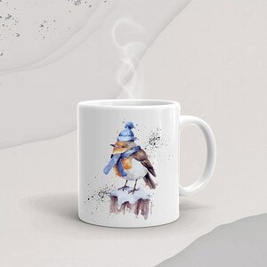 May include: White ceramic mug with a watercolor illustration of a robin wearing a blue hat and scarf, perched on a snow-covered branch. The mug has a black handle.